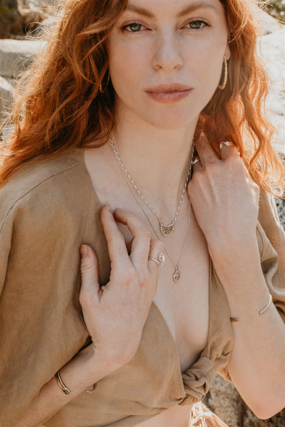 Woman with long hair wearing a beige top and layered necklaces outdoors.