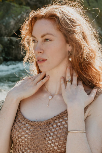 Woman touching her neck, wearing a necklace and ring, with a natural background.
