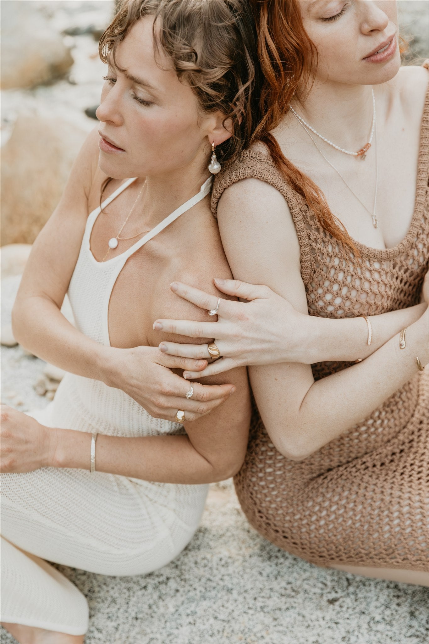 Two women sitting close together on a beach, embracing each other wearing jewelry
