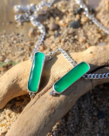 Necklace with green pendant on a piece of driftwood on a beach.