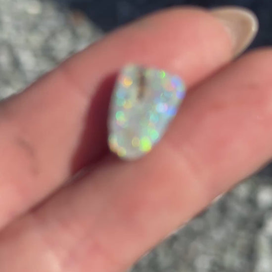 a video close-up of a opal stone with several different colors, held in a hand with a rocky background.