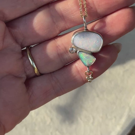 a close-up video of a person hand with an opal necklace. necklace has two opals and a moonstone, a small charm hangs below
